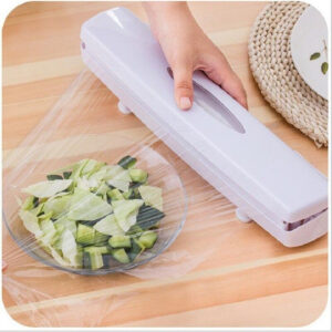 Dispensador de film plástico blanco en uso. Mano presionando herramienta para cubrir bowl de vidrio con ensalada con lámina transparente sobre mesa de madera.