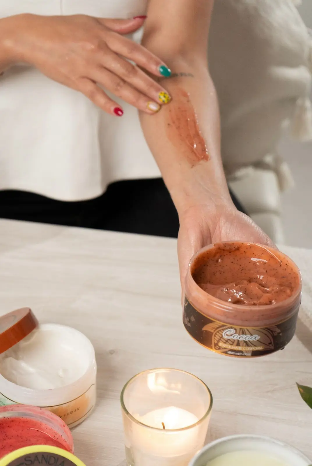 Mano aplicando exfoliante corporal de cocoa sobre el brazo. Frasco abierto a la vista.