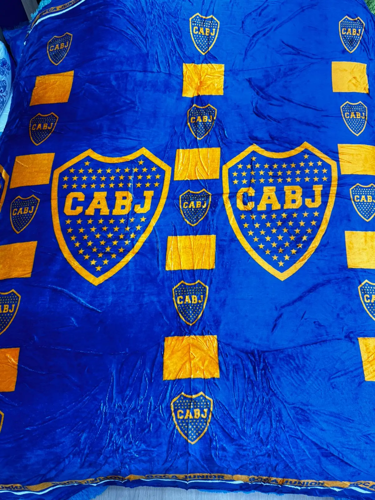 Cobija deportiva azul y dorada del CABJ, textura suave, diseño repetido con escudos y barras.
