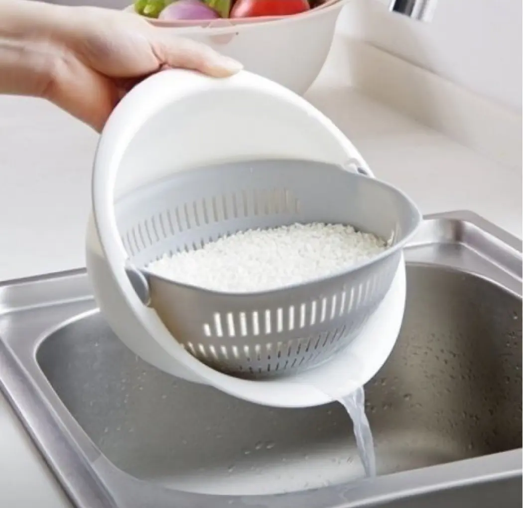 Colador giratorio blanco y gris con arroz y agua escurriendo en fregadero de acero inoxidable.