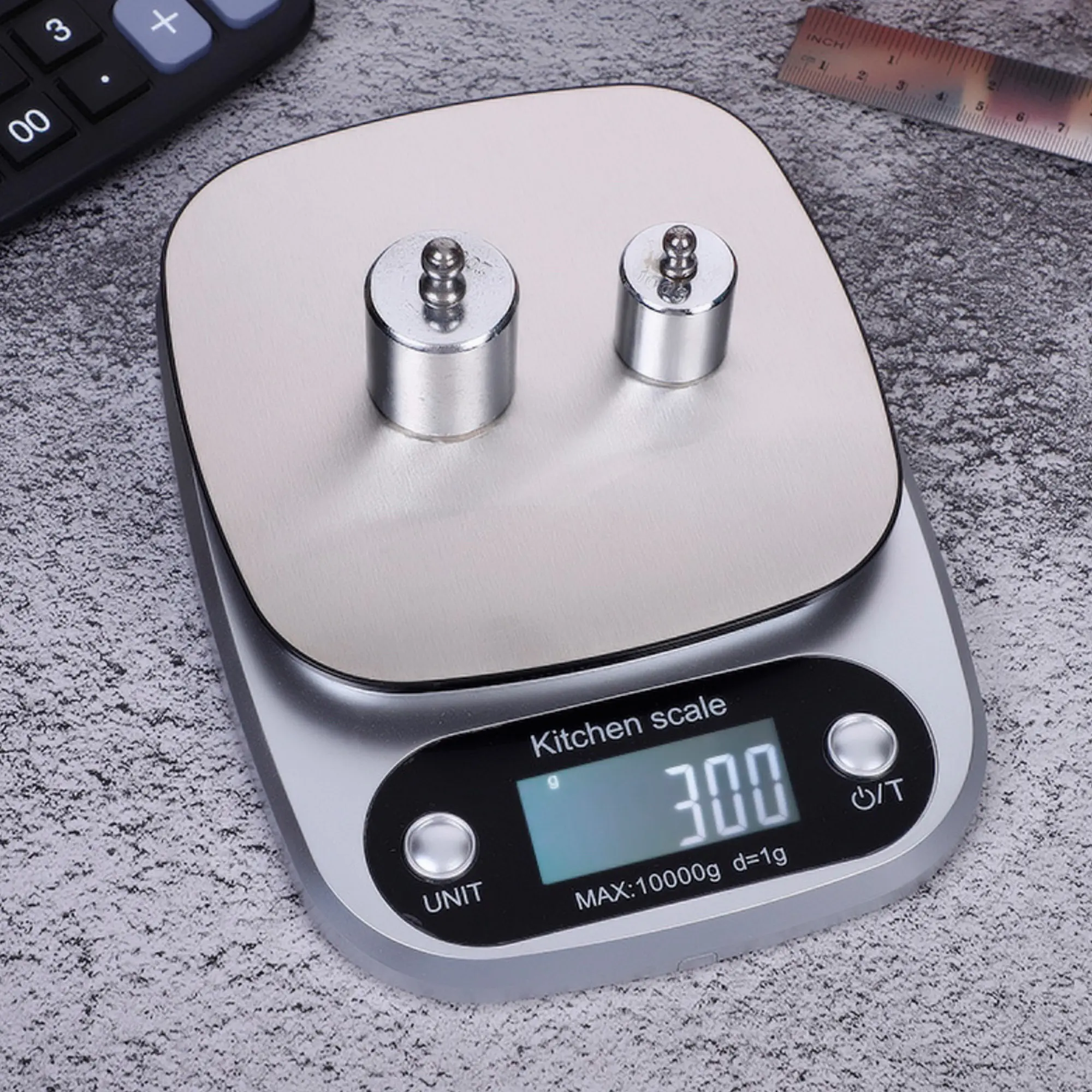 Balanza gramera digital plateada con dos pesas metálicas, muestra 300g. Ideal para cocina.