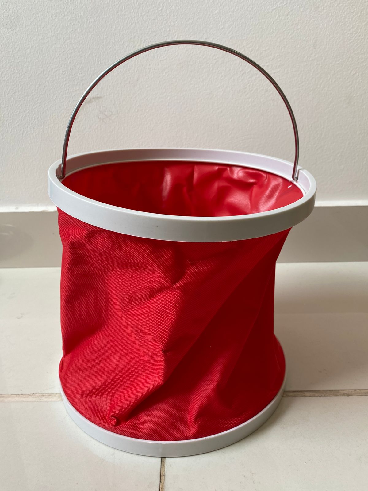 Balde de agua plegable rojo con asa metálica y borde blanco, colocado sobre baldosas.
