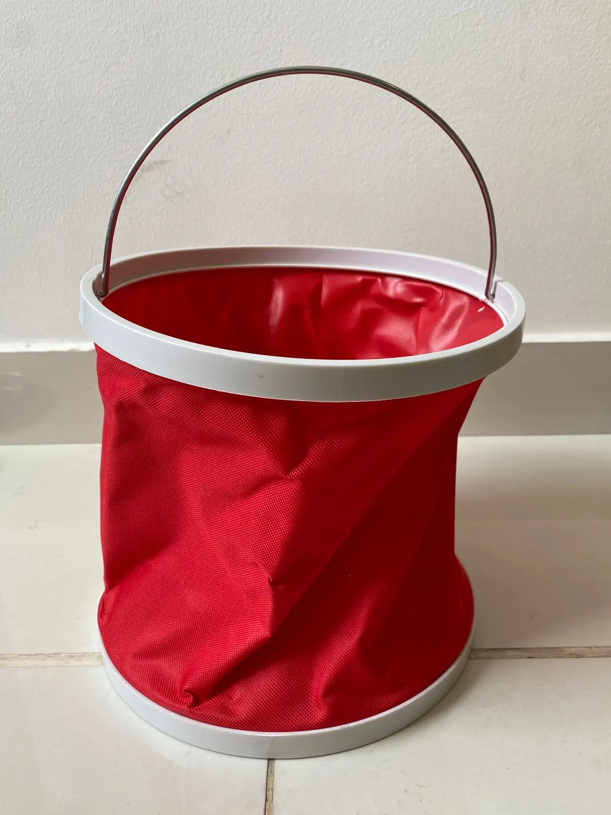 Balde de agua plegable rojo con asa metálica y borde blanco, colocado sobre baldosas.
