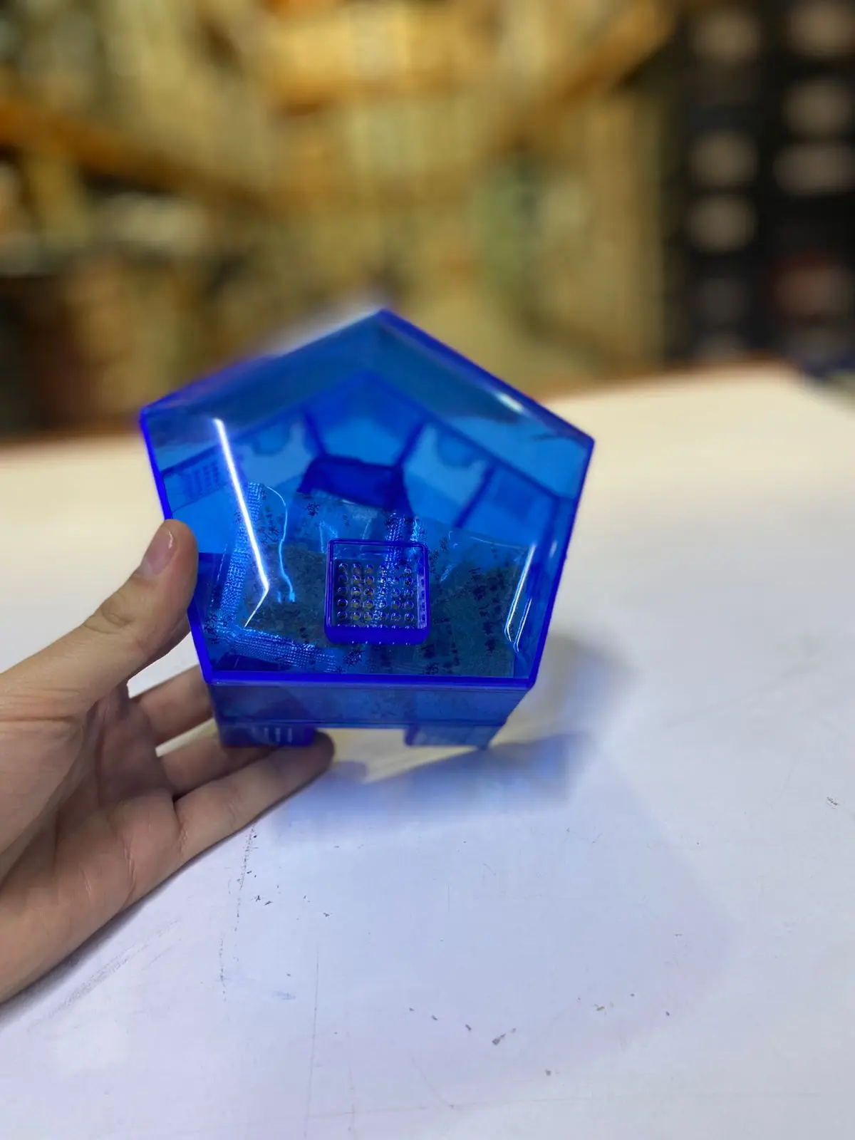 Trampa para cucarachas azul transparente en forma pentagonal, sujeta por mano. Contiene pequeñas bolsas y compartimento central cuadriculado.