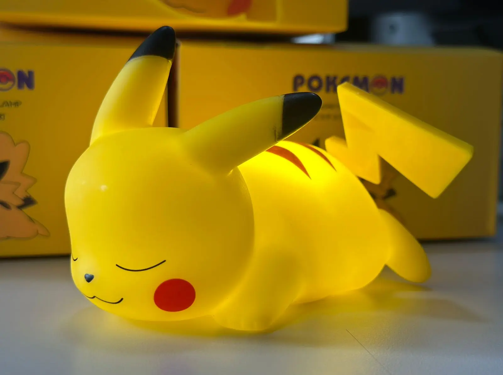 Lámpara LED Pikachu dormido, diseño infantil amarillo, con caja Pokémon al fondo.