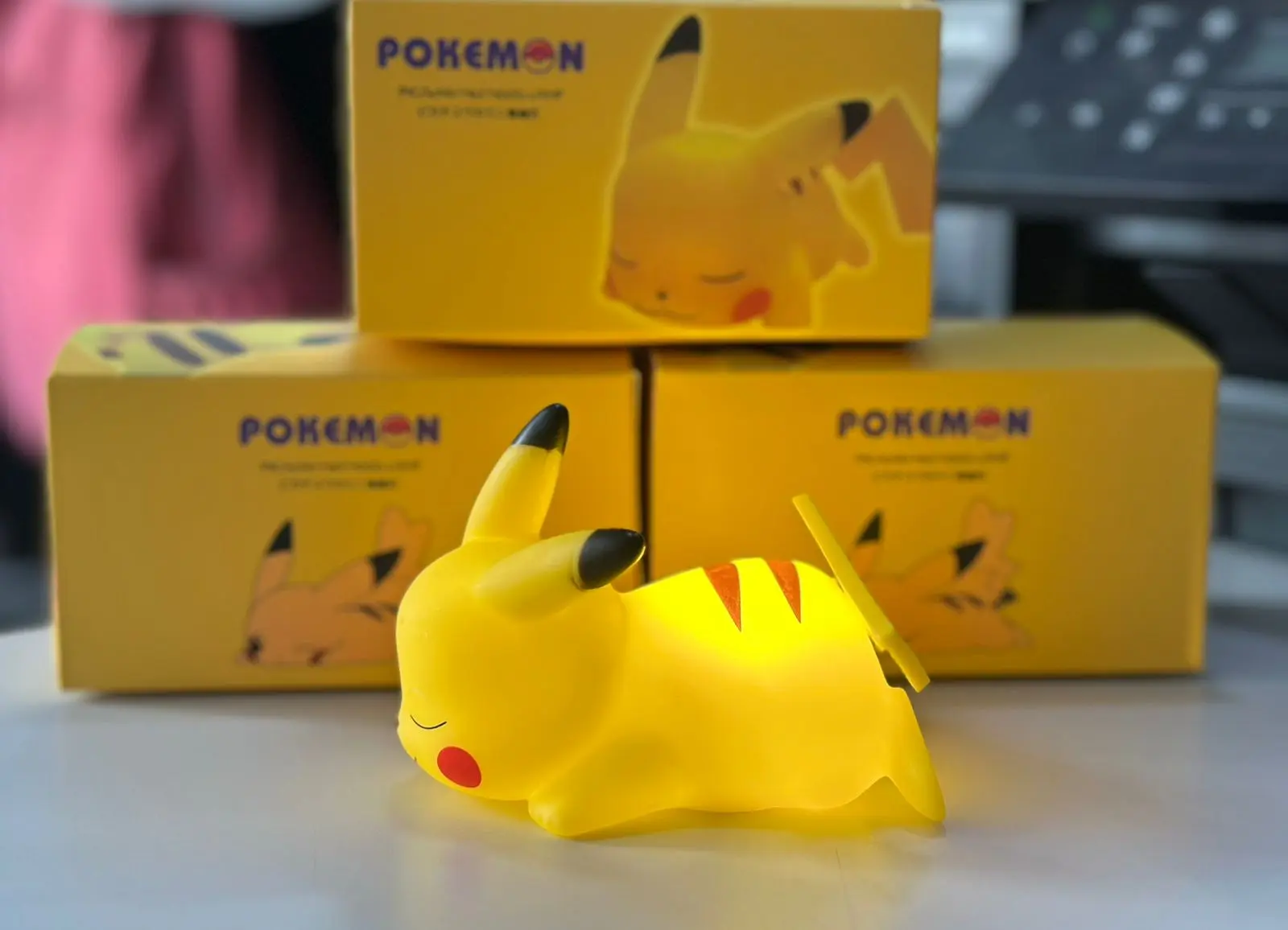 Lámpara LED de Pikachu durmiendo, diseño infantil, color amarillo brillante, con cajas de Pokémon al fondo.