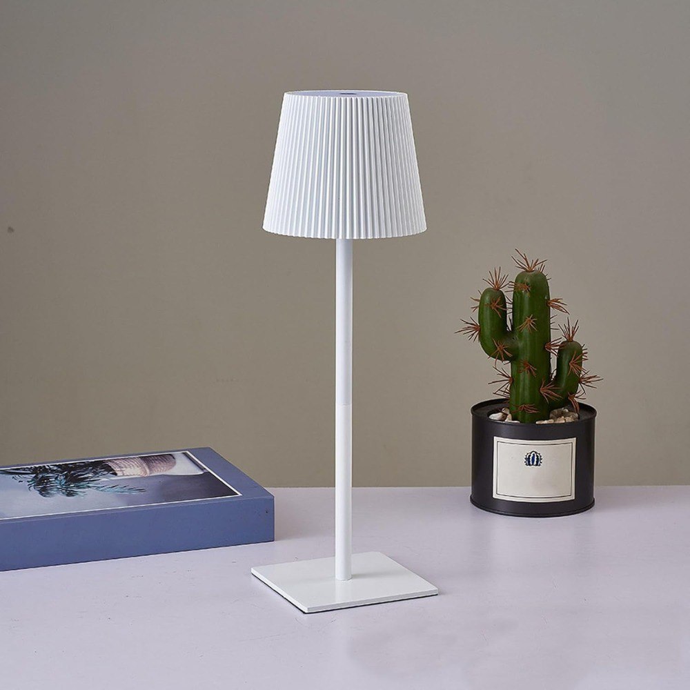 Lámpara LED minimalista blanca, metálica, recargable, con pantalla plisada, junto a cactus y libro.
