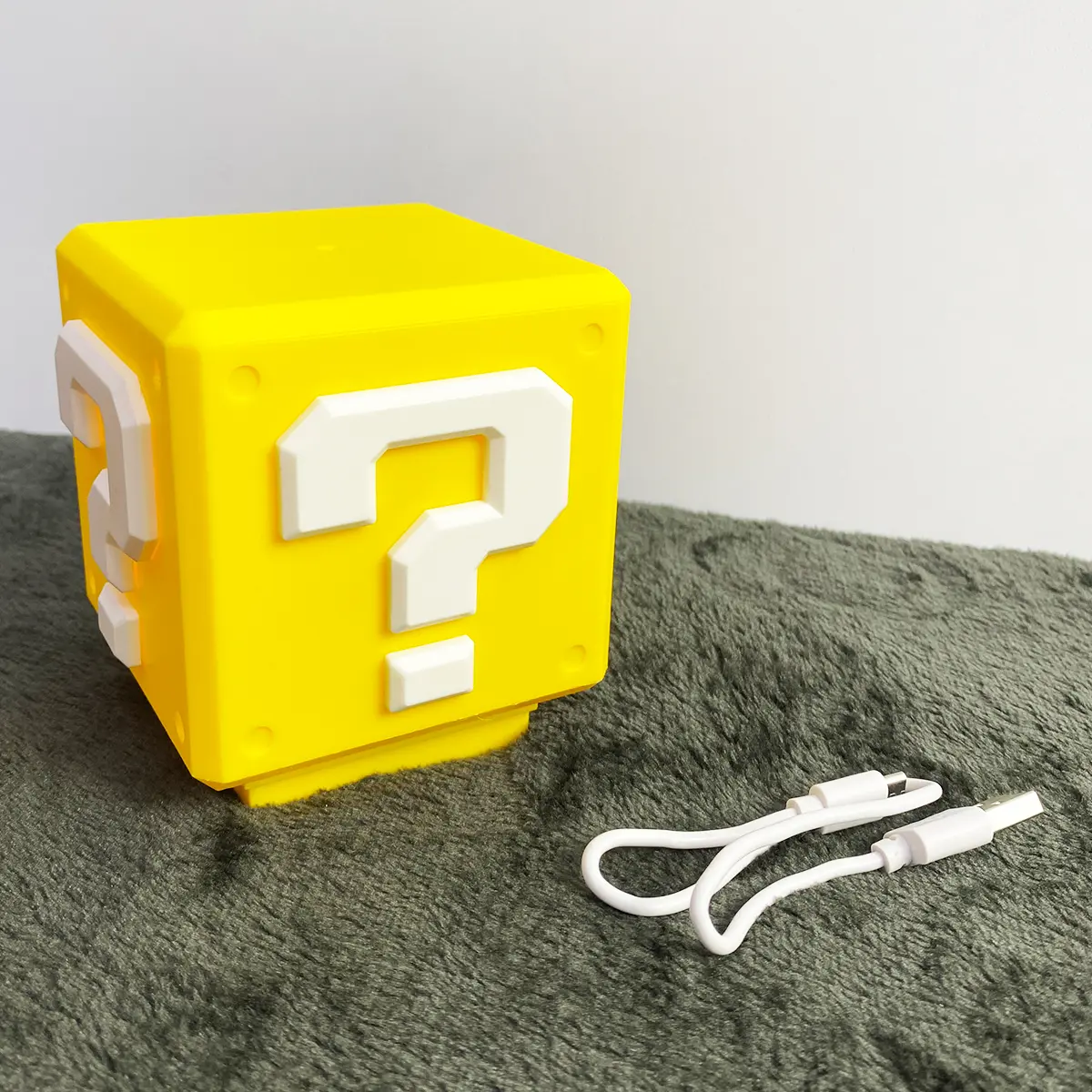 Lámpara en forma de bloque de interrogación Super Mario amarillo, con luces LED y cable USB, sobre superficie texturizada gris.