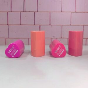 Cuatro rubores en barra, dos en forma de corazón rosa, uno cilíndrico naranja y otro cilíndrico rosa, sobre superficie blanca y fondo de ladrillos rosados.