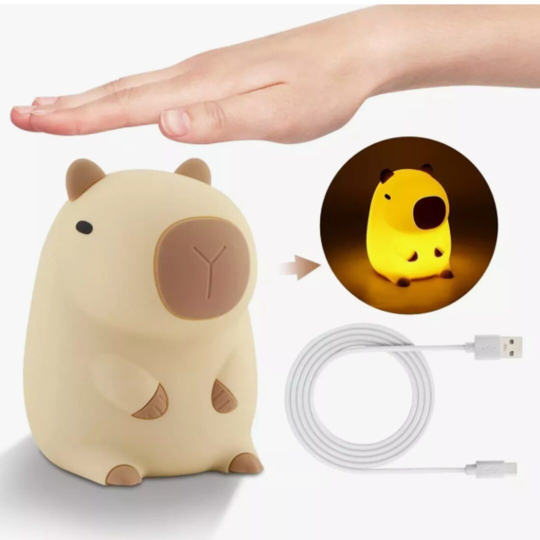 Lámpara de silicona con forma de capibara color beige, incluye cable de carga USB y muestra el efecto de luz cálida encendida junto a una mano humana.