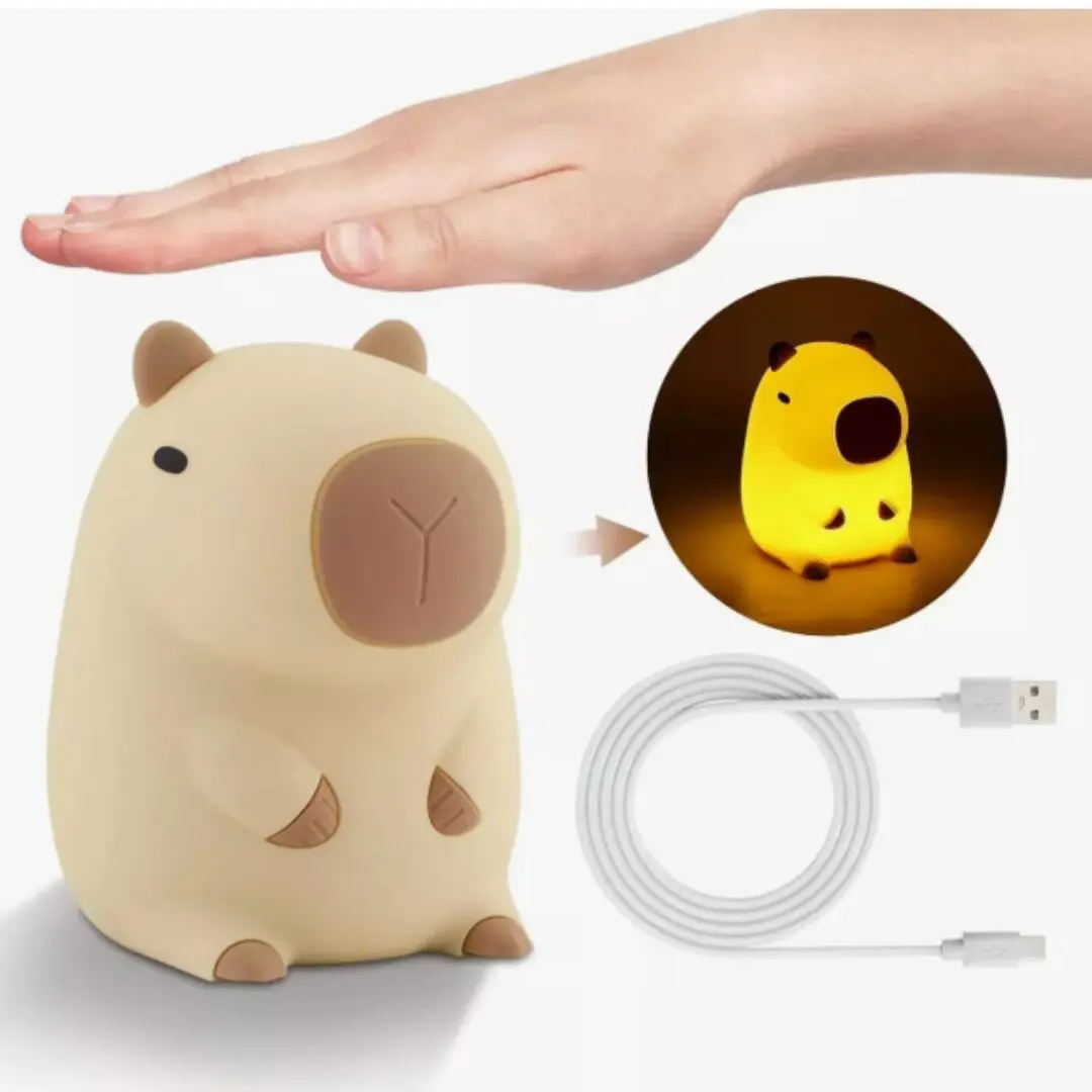 Lámpara de silicona con forma de capibara color beige, incluye cable de carga USB y muestra el efecto de luz cálida encendida junto a una mano humana.