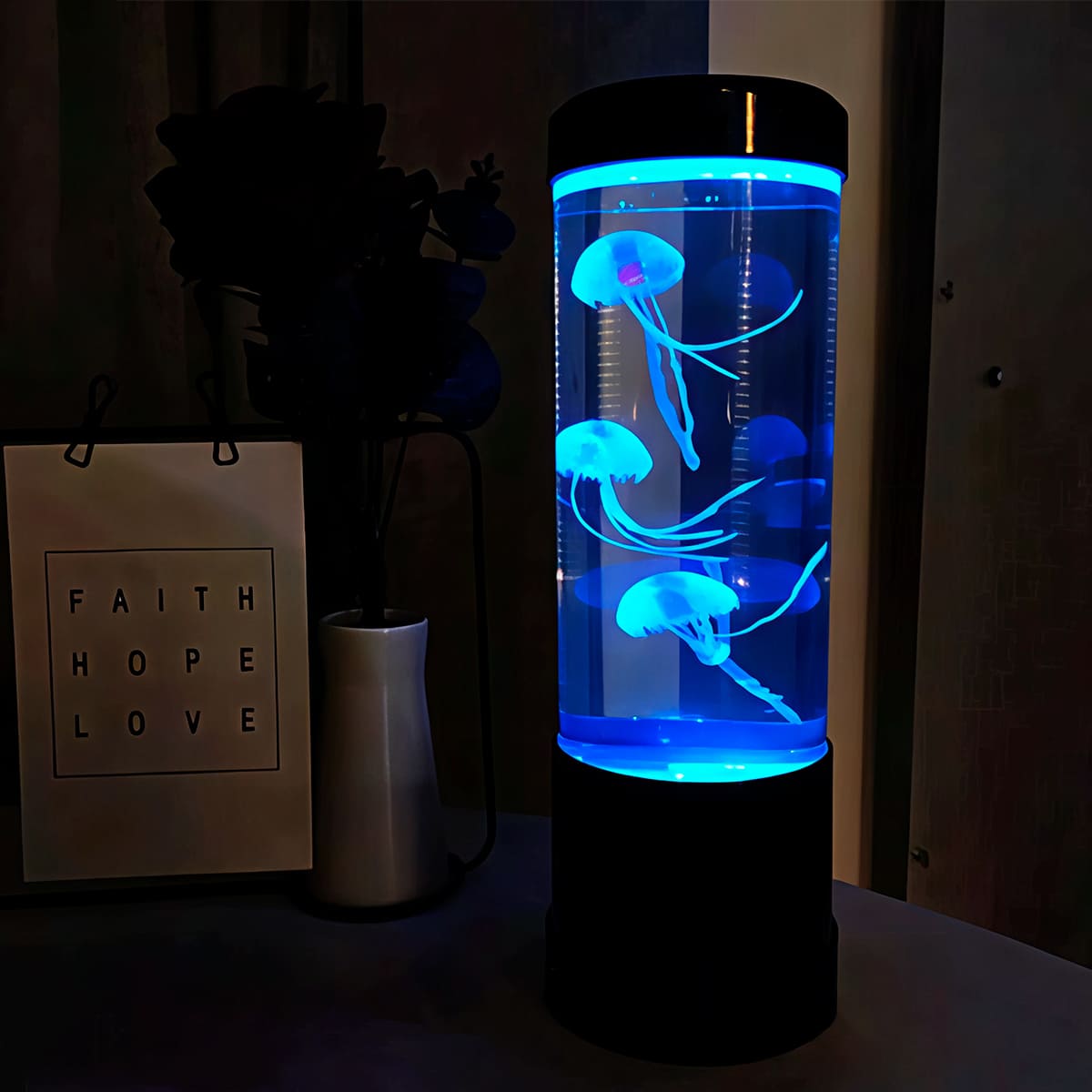 Lámpara LED cilíndrica con tres medusas artificiales que flotan en agua iluminada con luz azul intensa, sobre base negra, junto a letrero decorativo.