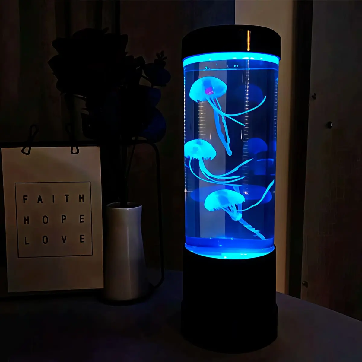 Lámpara LED cilíndrica con tres medusas artificiales que flotan en agua iluminada con luz azul intensa, sobre base negra, junto a letrero decorativo.
