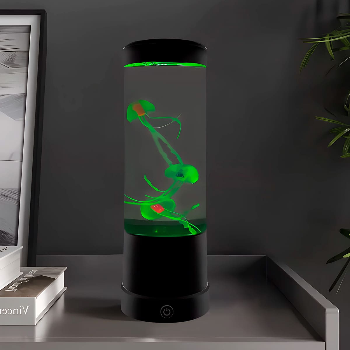 Lámpara LED cilíndrica con medusas artificiales flotantes iluminadas en color verde, base negra con interruptor táctil, ubicada sobre una mesa gris.
