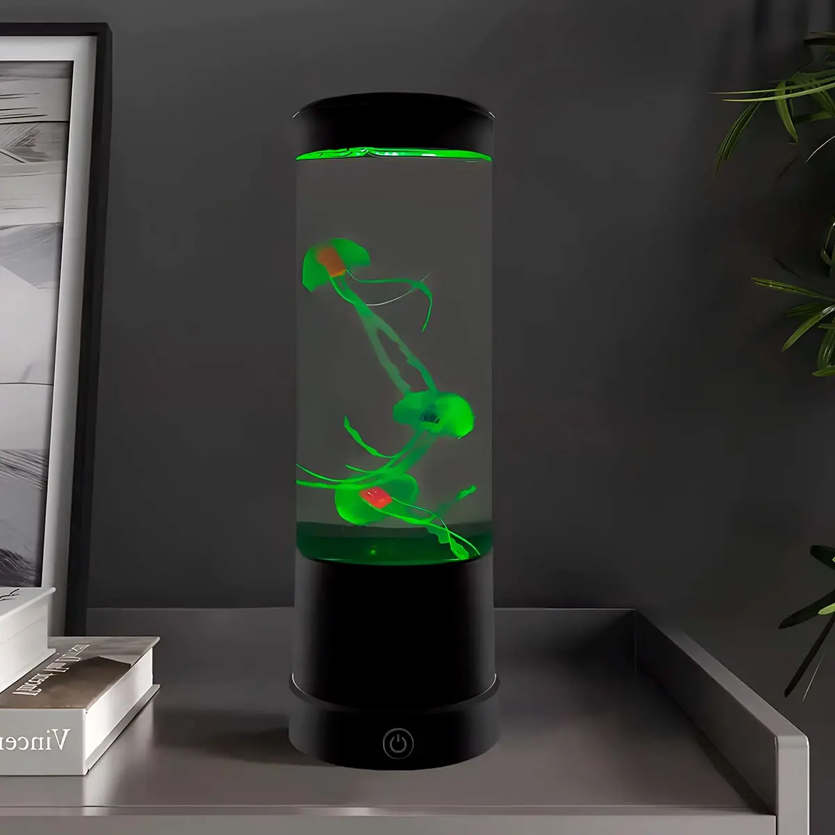 Lámpara LED cilíndrica con medusas artificiales flotantes iluminadas en color verde, base negra con interruptor táctil, ubicada sobre una mesa gris.