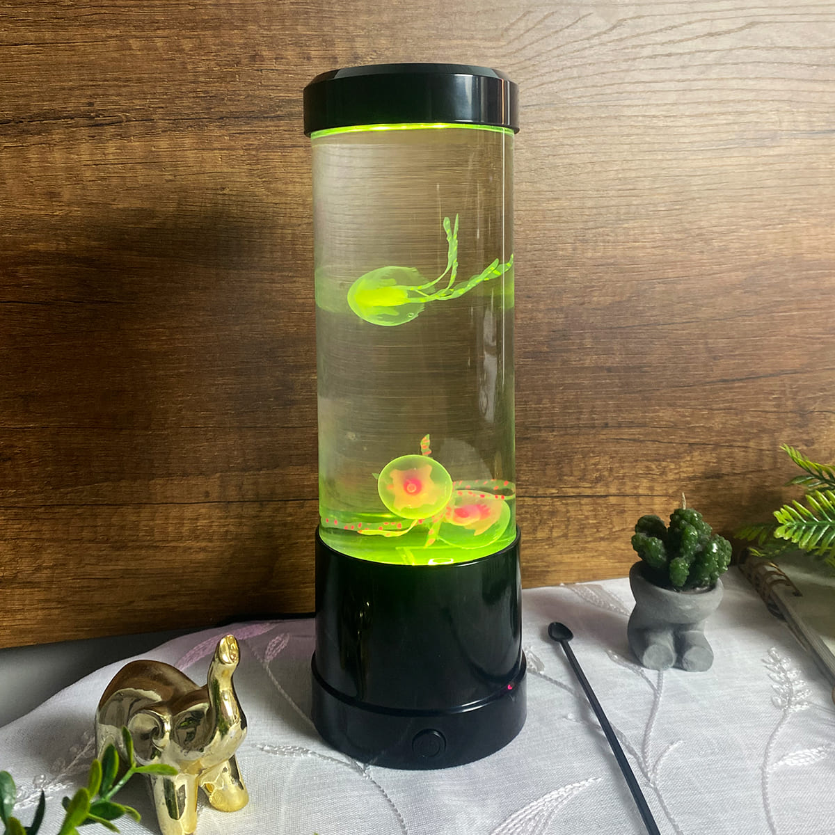 Lámpara LED cilíndrica con dos medusas de silicona en su interior, iluminación verde, base negra y accesorios decorativos sobre mesa blanca.