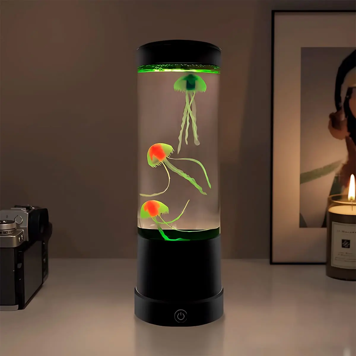 Lámpara LED cilíndrica con tres medusas artificiales multicolor flotando, base negra con interruptor frontal, sobre superficie blanca junto a una cámara.