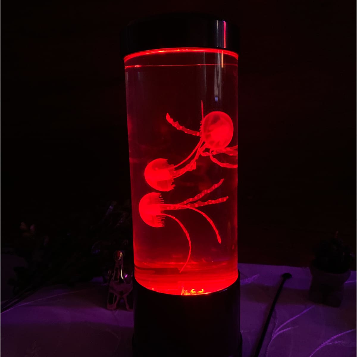 Lámpara LED cilíndrica con tres medusas artificiales flotando en su interior, iluminada con luz de tono rojo intenso sobre una base negra.