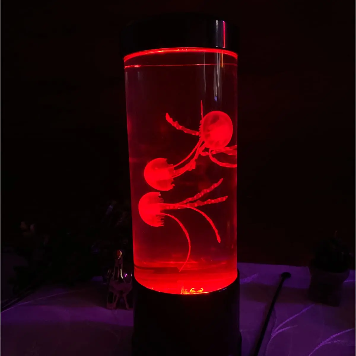 Lámpara LED cilíndrica con tres medusas artificiales flotando en su interior, iluminada con luz de tono rojo intenso sobre una base negra.