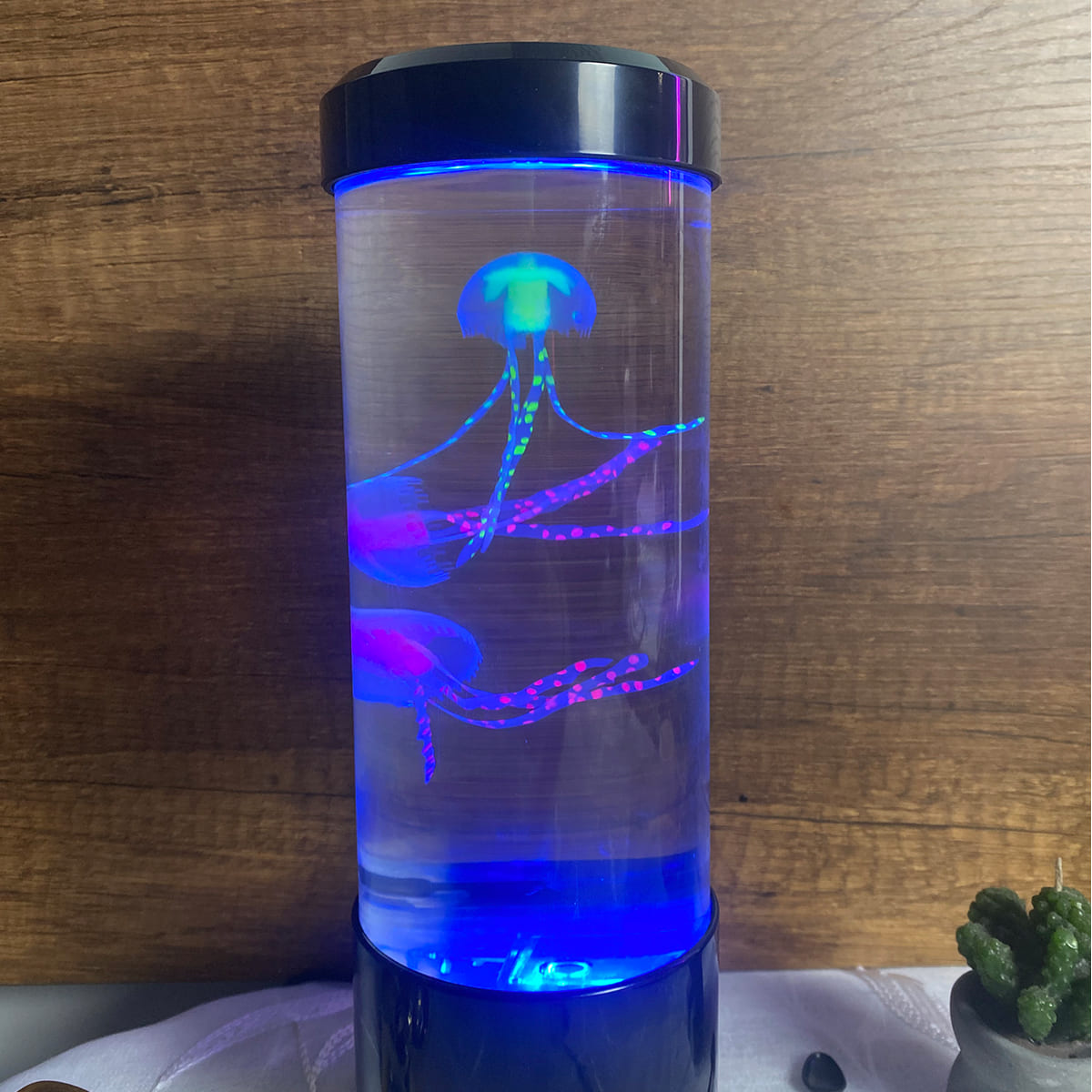 Lámpara LED cilíndrica con dos medusas artificiales que flotan y brillan en tonos azul, verde y fucsia sobre fondo de madera.