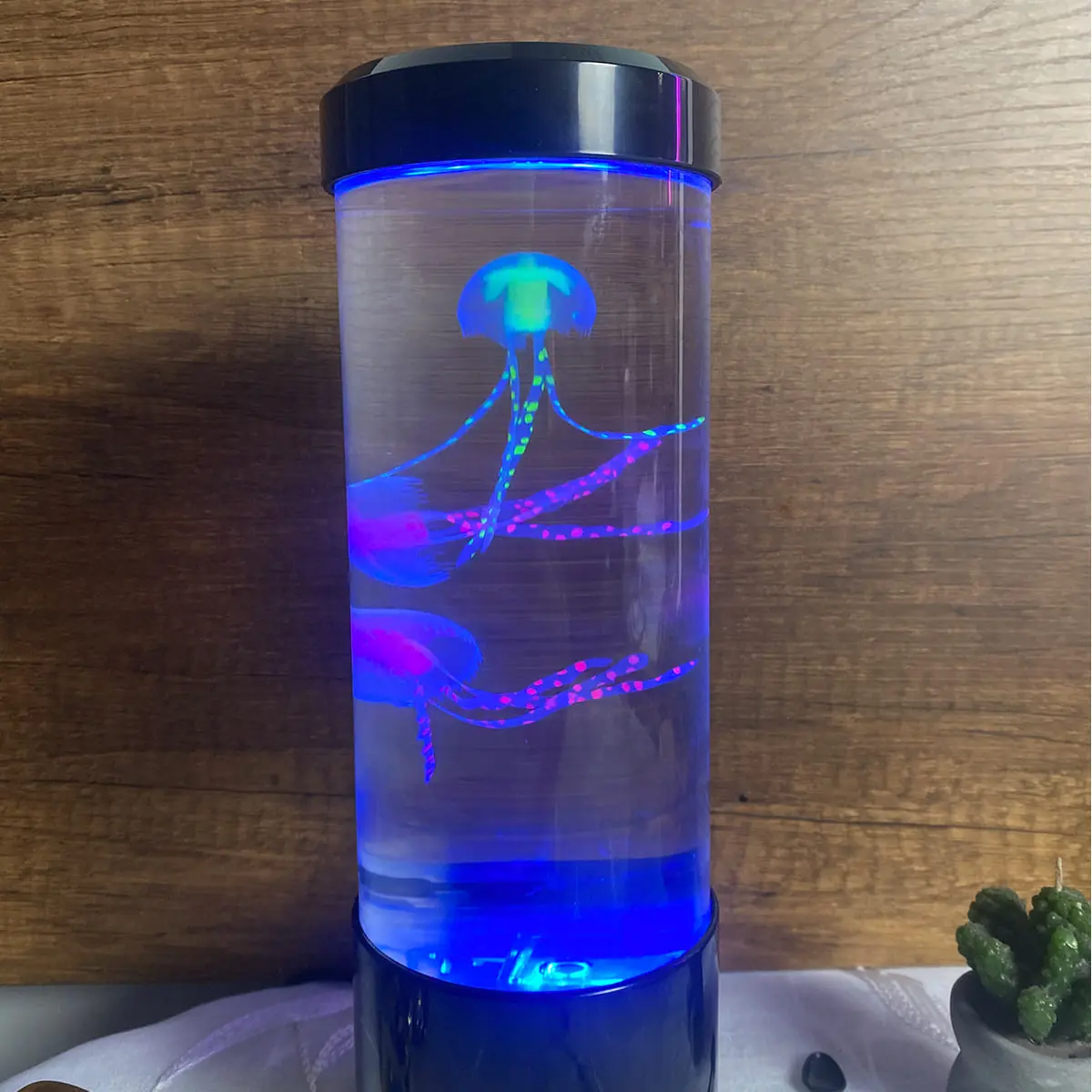 Lámpara LED cilíndrica con dos medusas artificiales que flotan y brillan en tonos azul, verde y fucsia sobre fondo de madera.
