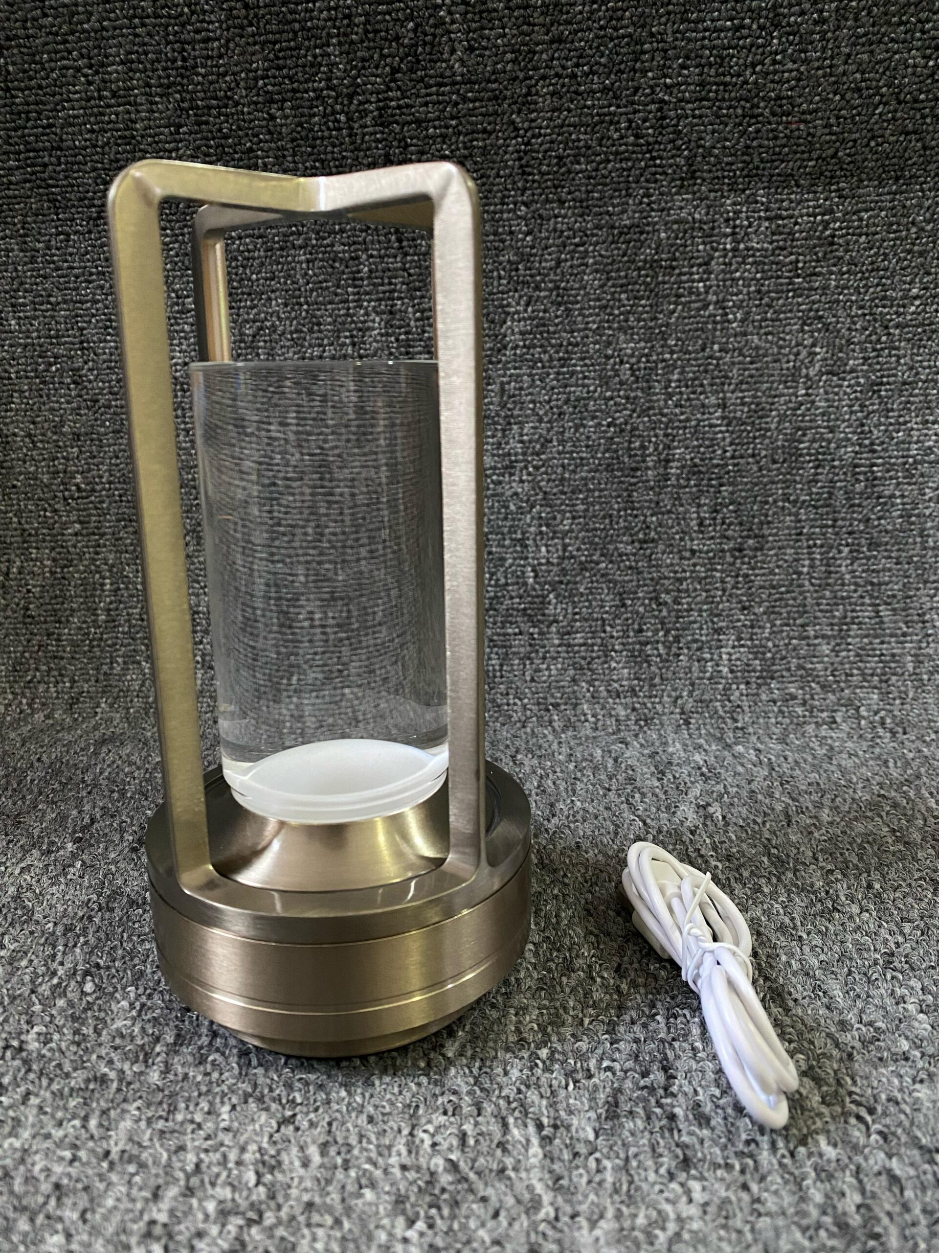 Lámpara de luz táctil metálica color bronce con cilindro de vidrio transparente y cable de carga blanco, sobre superficie de textura gris.