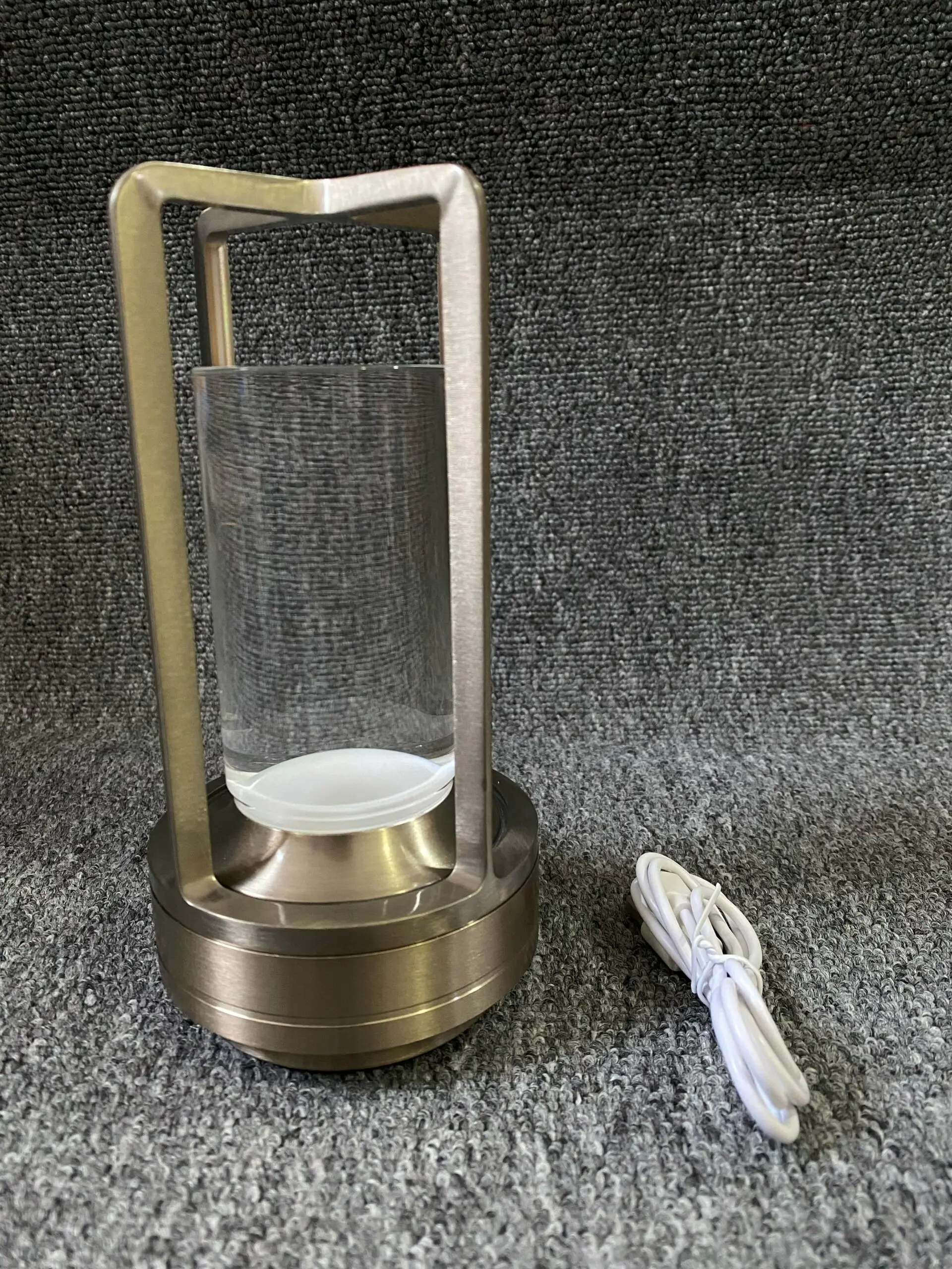 Lámpara de luz táctil metálica color bronce con cilindro de vidrio transparente y cable de carga blanco, sobre superficie de textura gris.