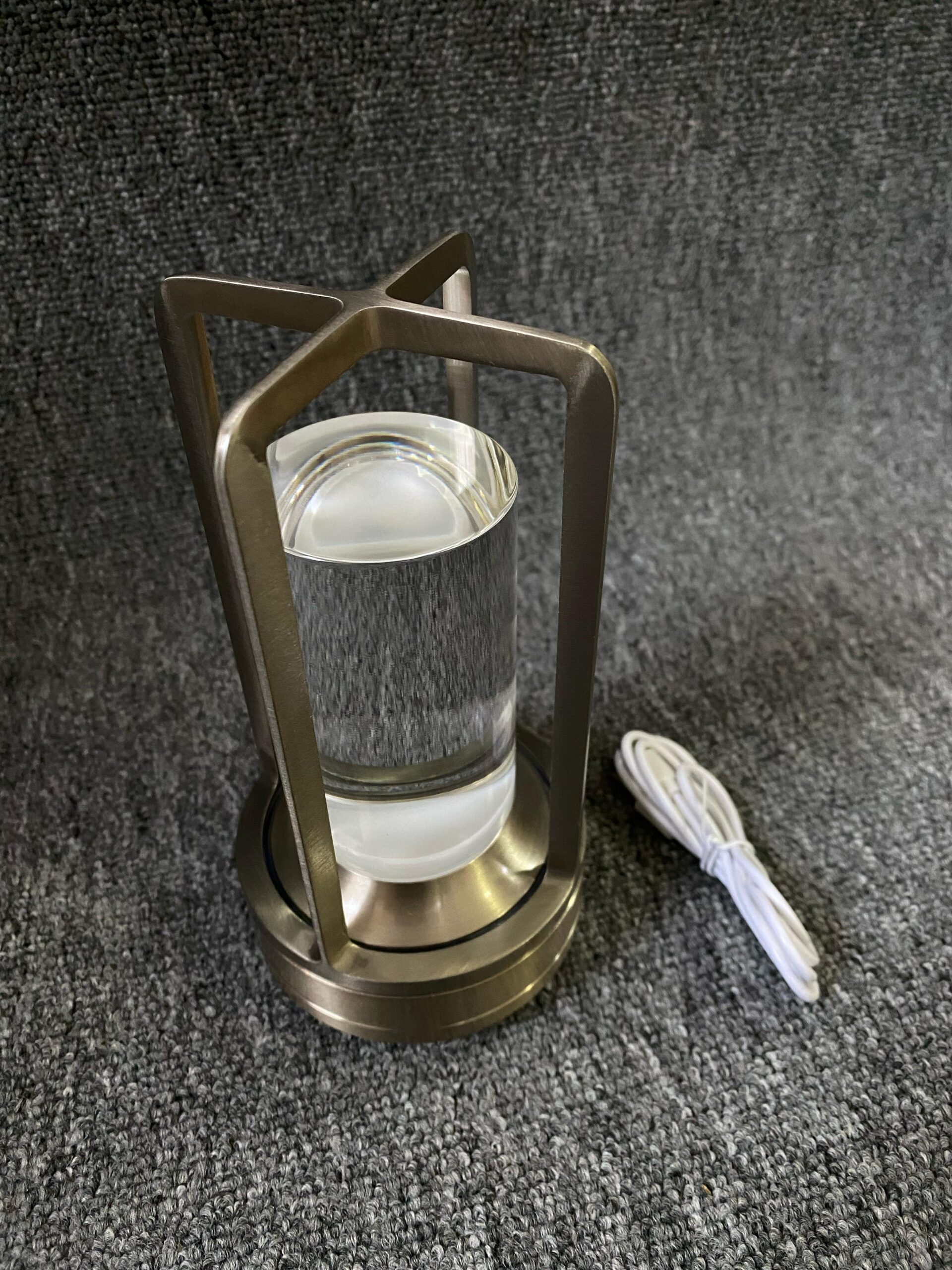 Lámpara de luz táctil con estructura metálica color bronce, difusor de vidrio cilíndrico transparente y cable de carga blanco incluido sobre fondo gris.