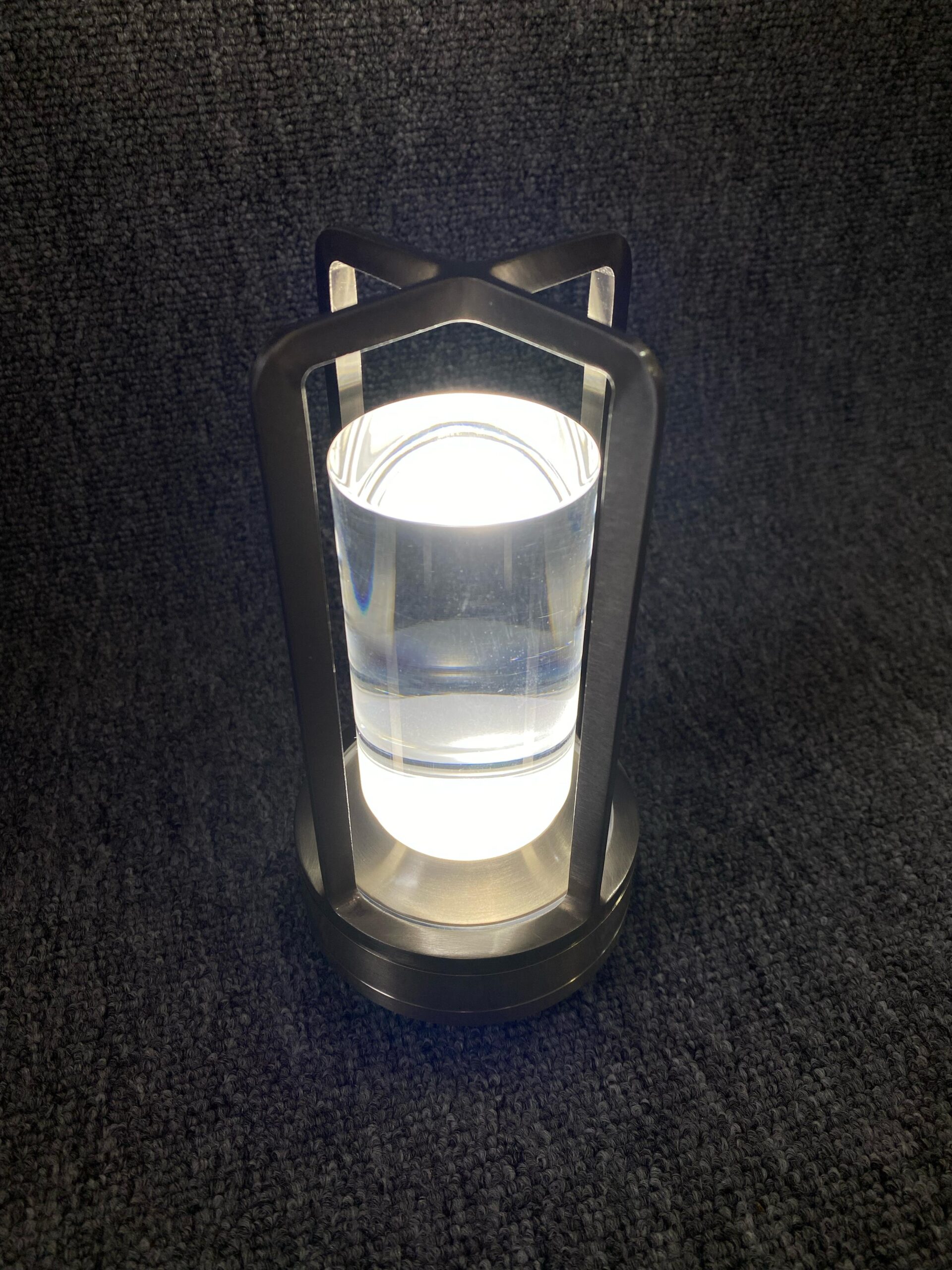 Lámpara de mesa táctil con base y estructura metálica color níquel, cuerpo cilíndrico de acrílico transparente y luz LED cálida encendida sobre fondo gris.