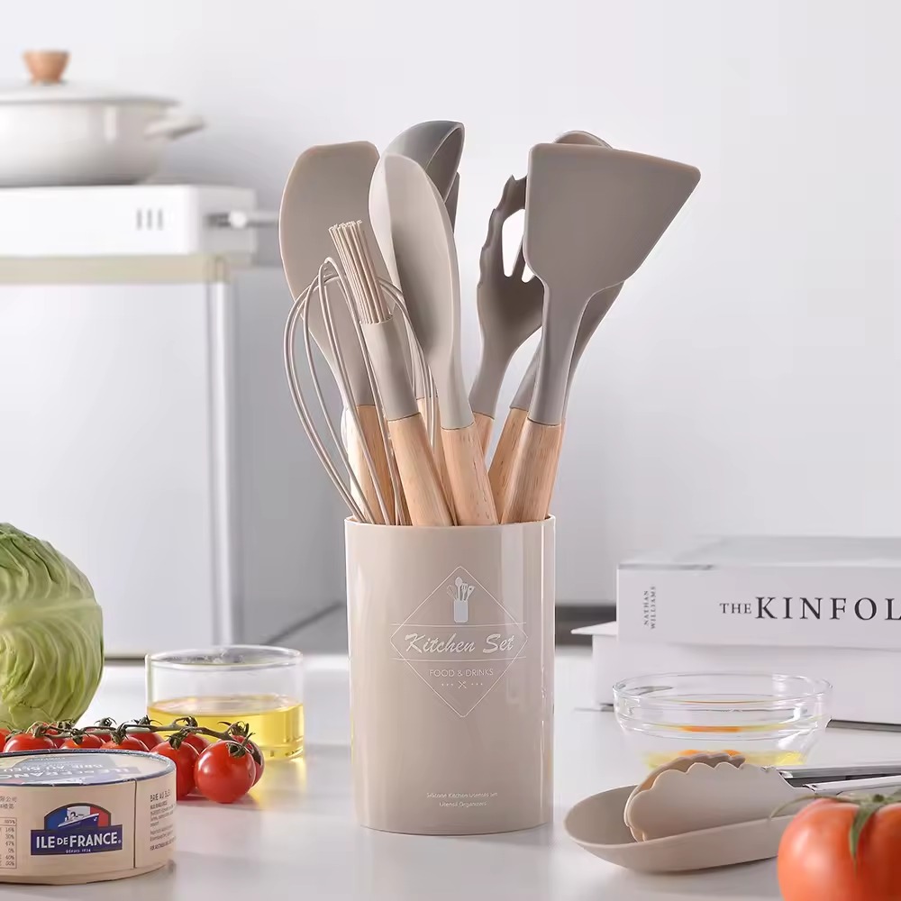 Set de 12 utensilios de cocina beige con mangos de madera, organizados en un recipiente cilíndrico a juego sobre una mesa junto a ingredientes frescos.
