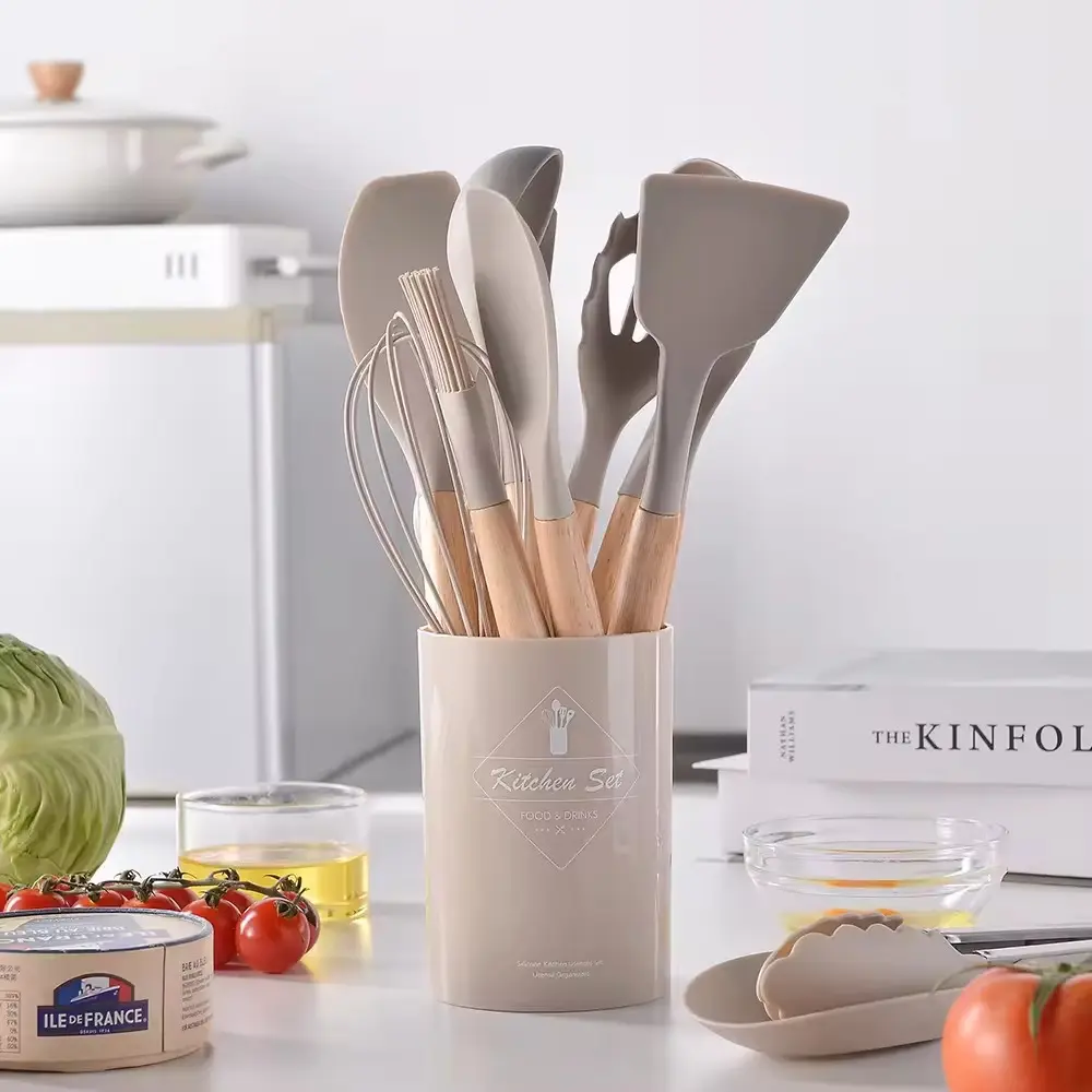 Set de 12 utensilios de cocina beige con mangos de madera, organizados en un recipiente cilíndrico a juego sobre una mesa junto a ingredientes frescos.