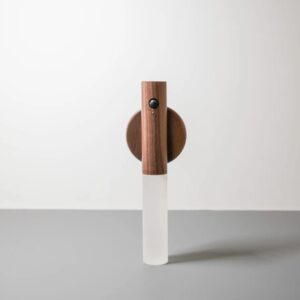 Lámpara portátil LED de pared con cuerpo cilíndrico en madera natural, difusor blanco esmerilado y sensor de movimiento frontal montada sobre base circular.