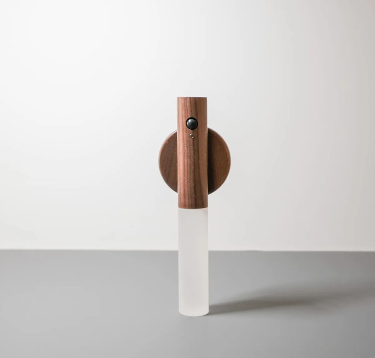 Lámpara portátil LED de pared con cuerpo cilíndrico en madera natural, difusor blanco esmerilado y sensor de movimiento frontal montada sobre base circular.