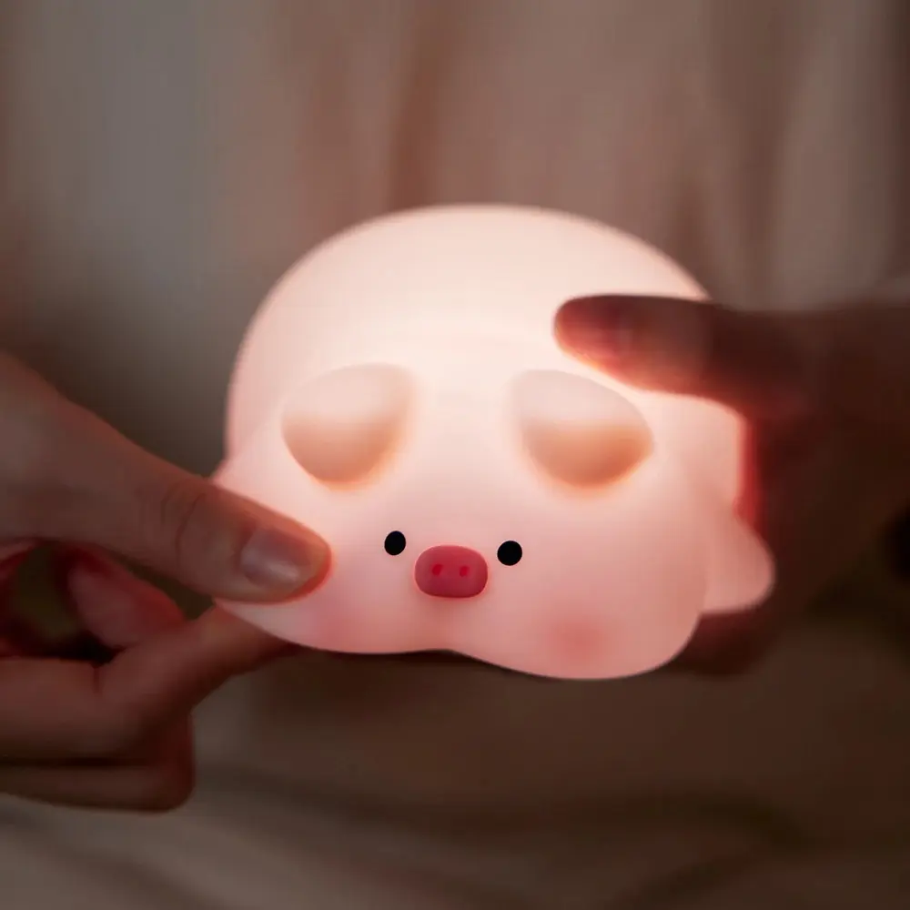 Lampara de goma rosa con forma de cerdito, iluminada, sostenida por manos.