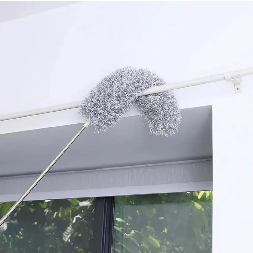 Plumero extensible de microfibra gris curvado limpiando el borde superior de una ventana con marco blanco.