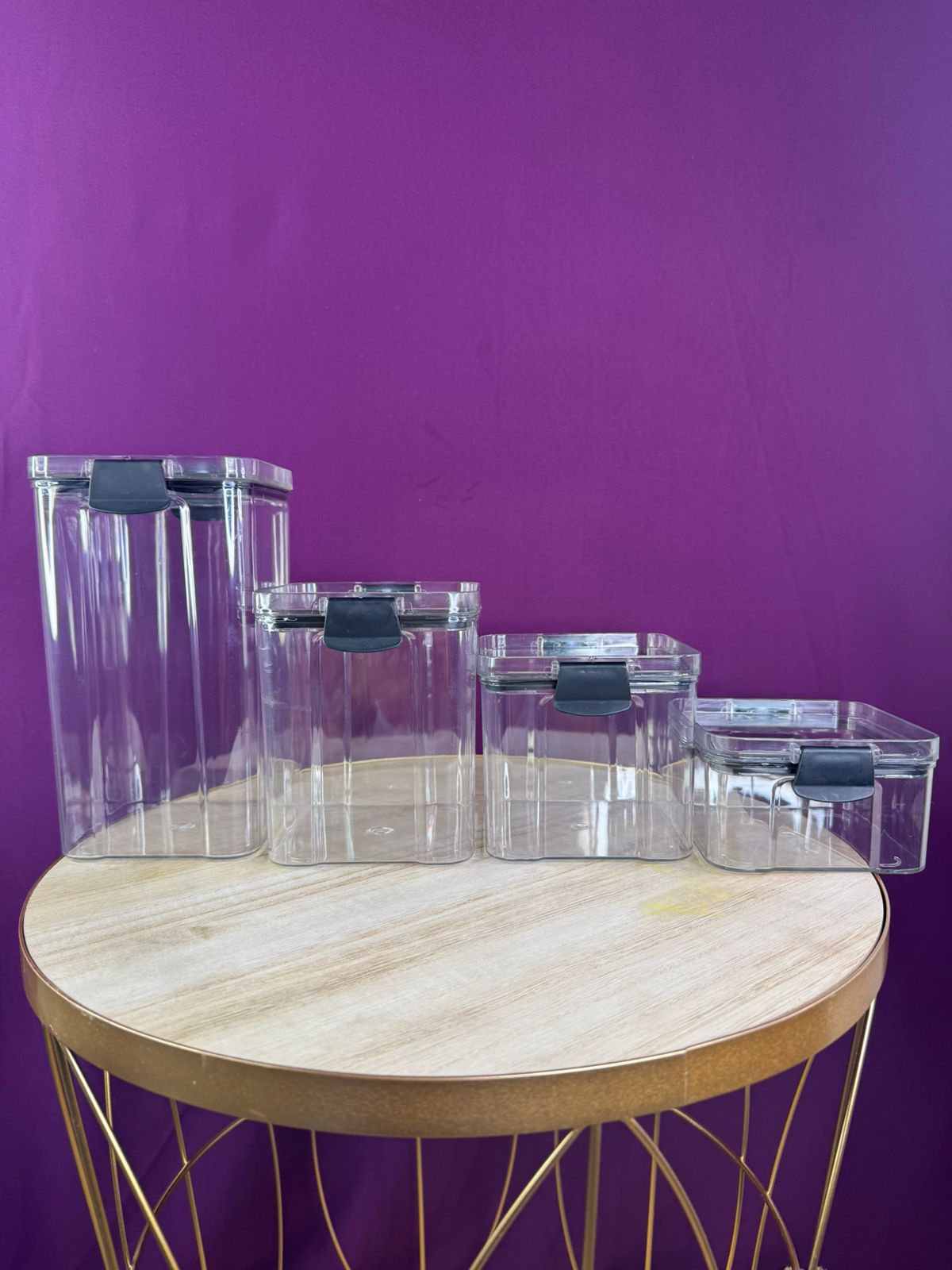 Cuatro organizadores de cocina rectangulares transparentes de plástico con tapa negra sobre mesa redonda dorada y fondo morado.
