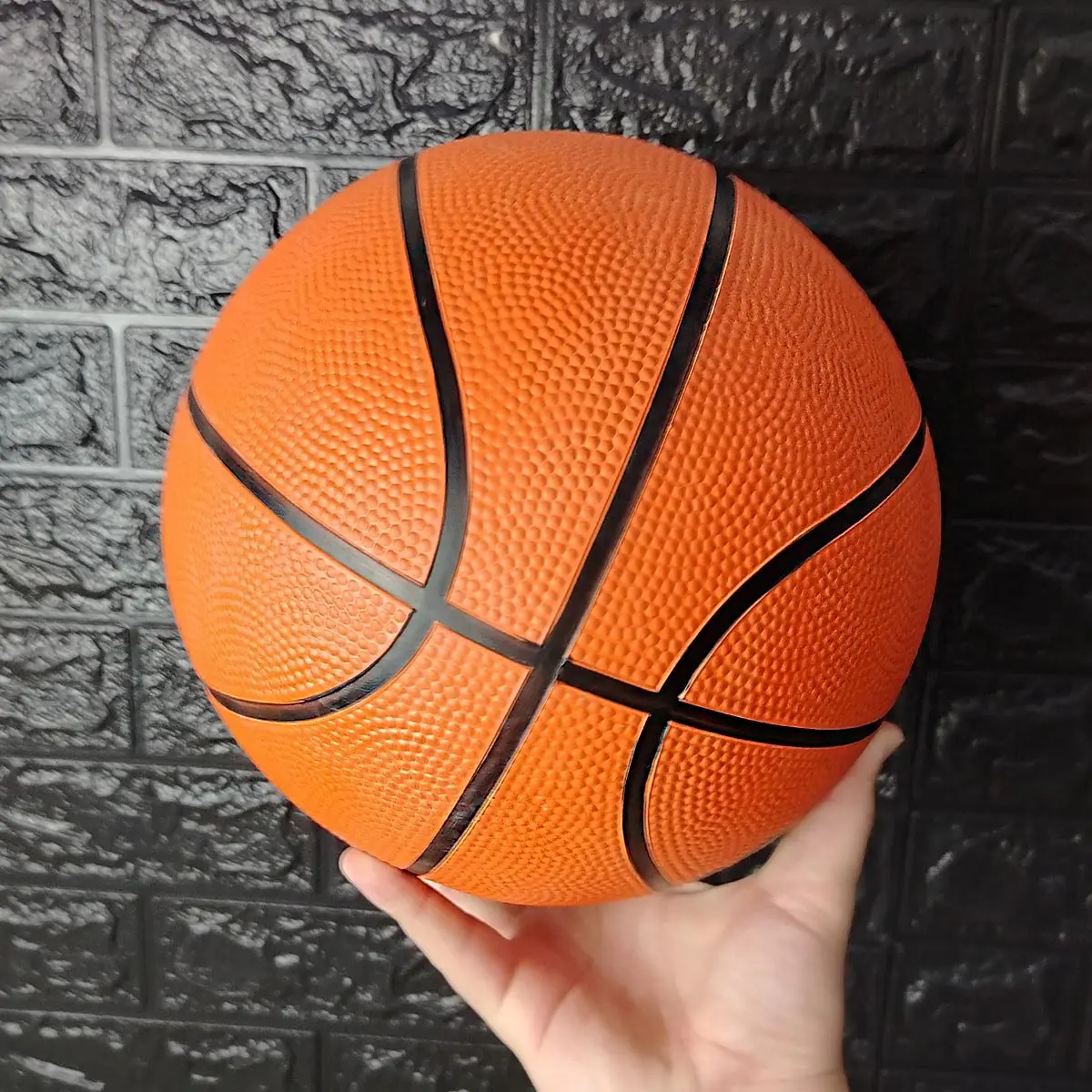 Balón de baloncesto naranja número 7, con textura rugosa, sostenido en mano contra pared de ladrillo negro.