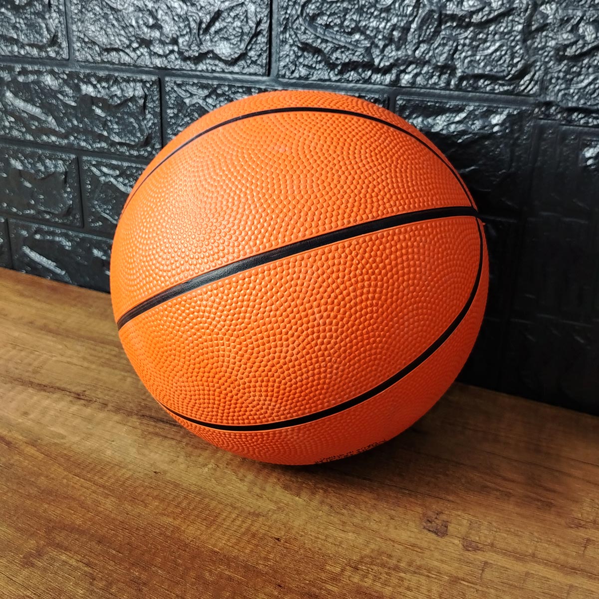 Balón de baloncesto naranja texturizado, tamaño 7, con líneas negras, sobre madera y fondo de ladrillo negro.