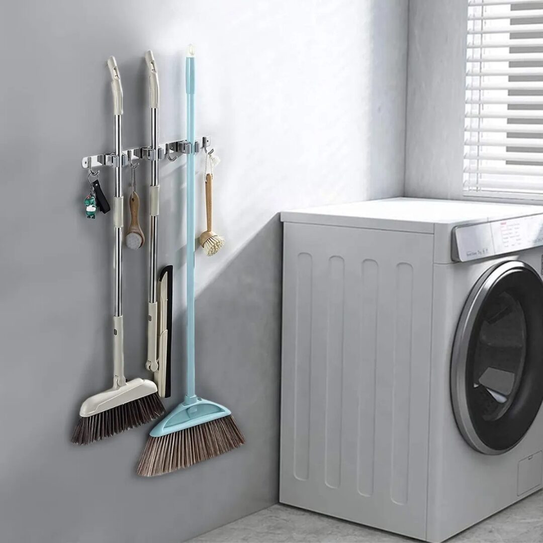 Soporte organizador de acero inoxidable para escobas y utensilios colgado en pared gris, junto a lavadora blanca.