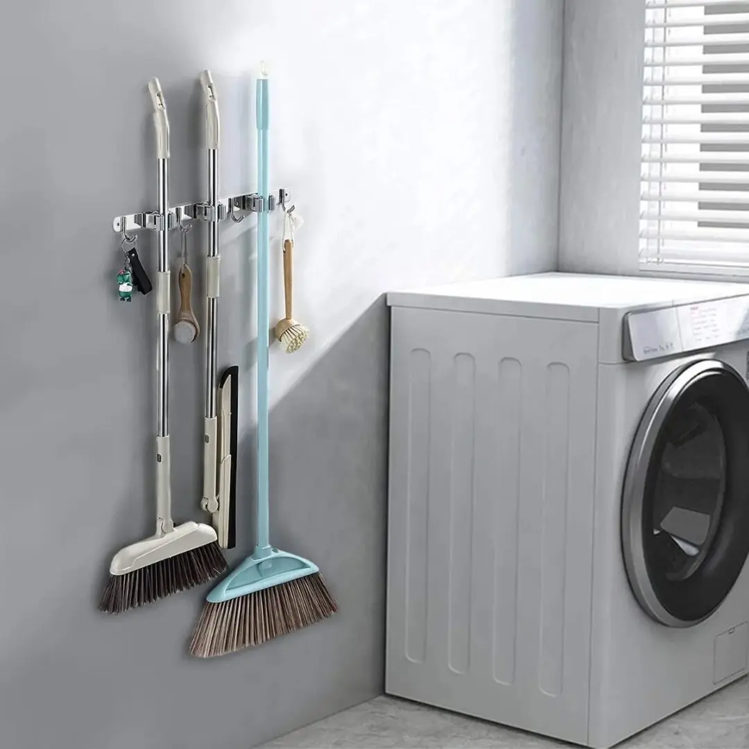 Soporte organizador de acero inoxidable para escobas y utensilios colgado en pared gris, junto a lavadora blanca.