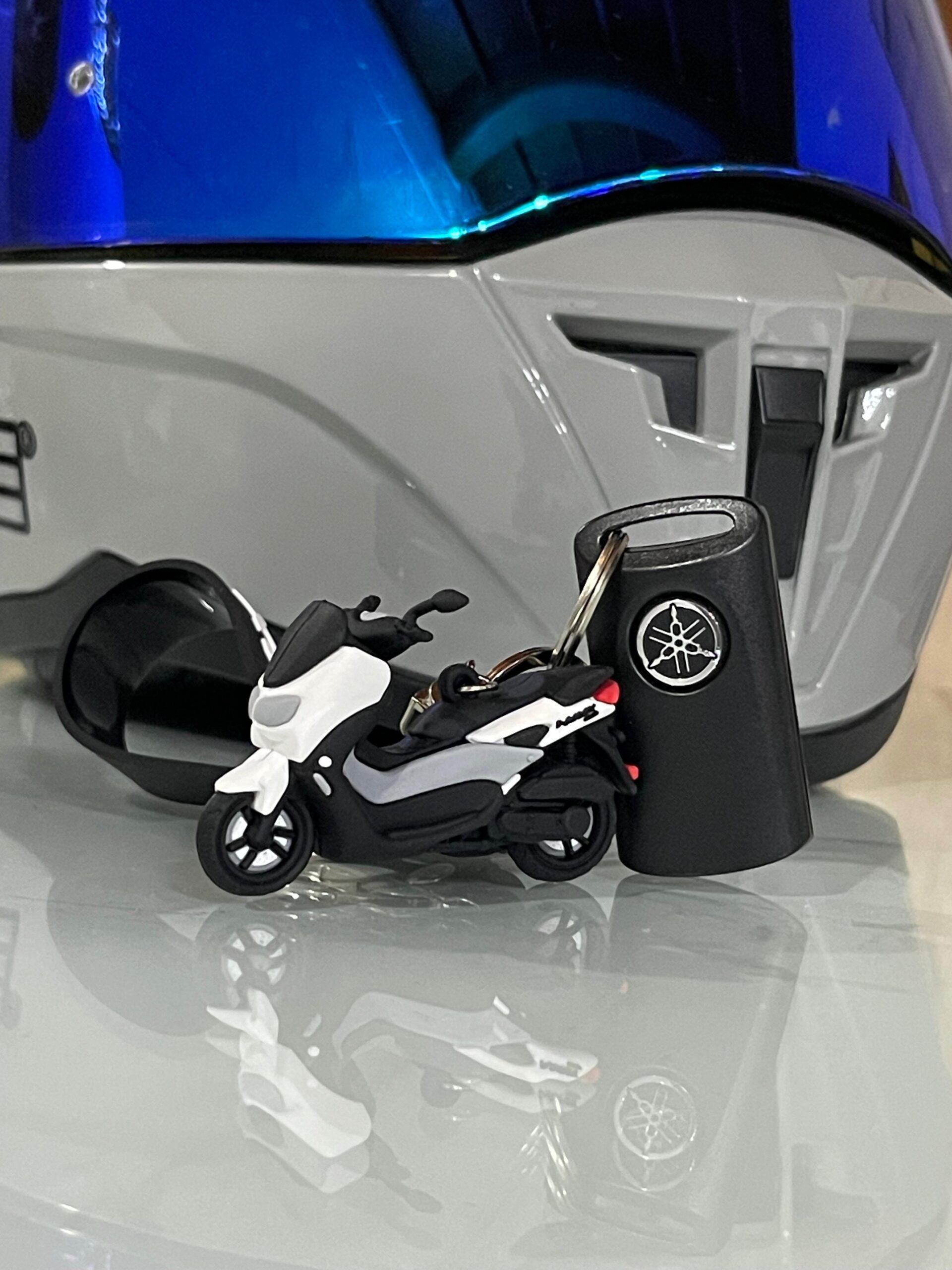 Llavero en 3D de moto Yamaha NMAX blanca y negra junto a su llave, sobre superficie reflectante.