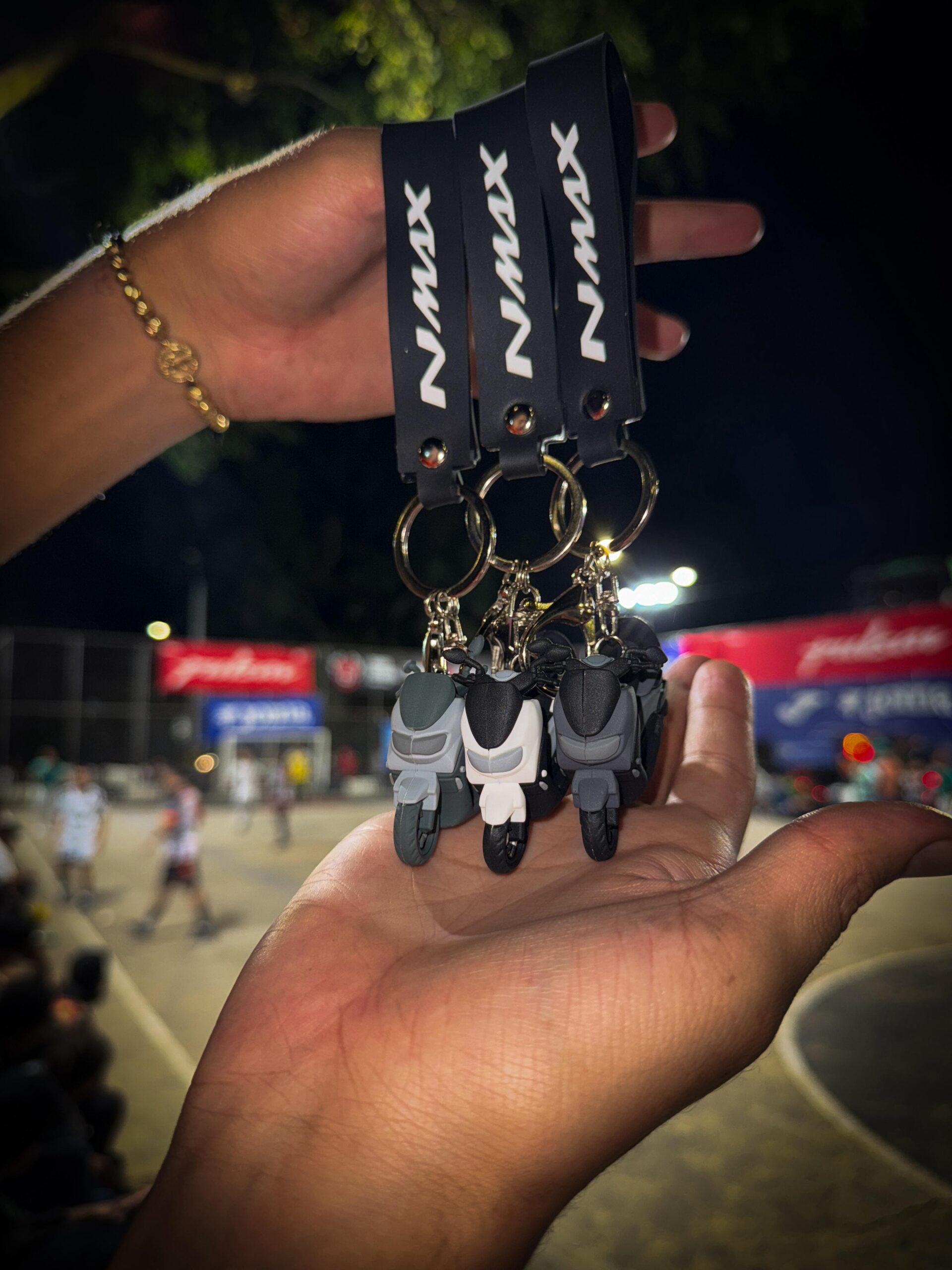 Llaveros 3D de moto NMAX grises y blancos, con correa negra, colgados en una mano.