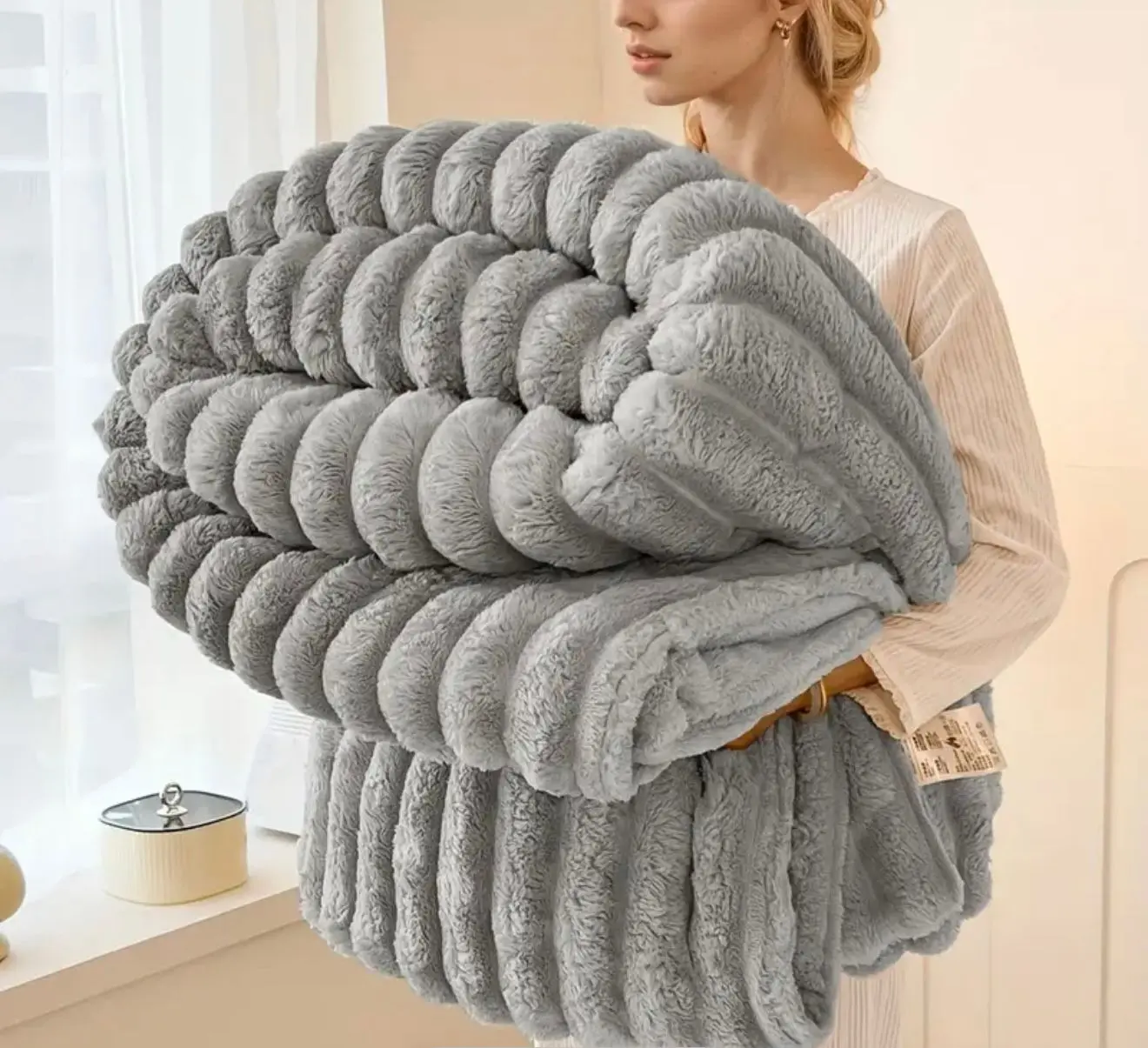 Manta polar felpa de conejo gris, textura acanalada, sostenida por una mujer al lado de una ventana.