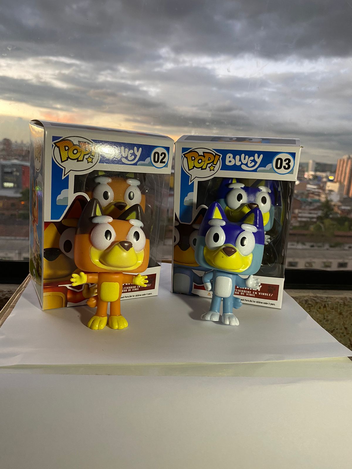 Figuras Funko Pop de Bluey y Bingo, naranja y azul, en sus cajas, sobre superficie blanca con fondo de atardecer.