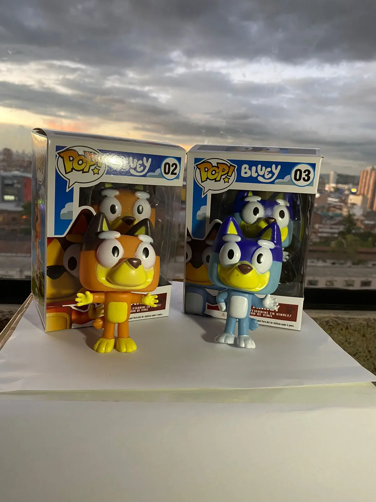 Figuras Funko Pop de Bluey y Bingo, naranja y azul, en sus cajas, sobre superficie blanca con fondo de atardecer.