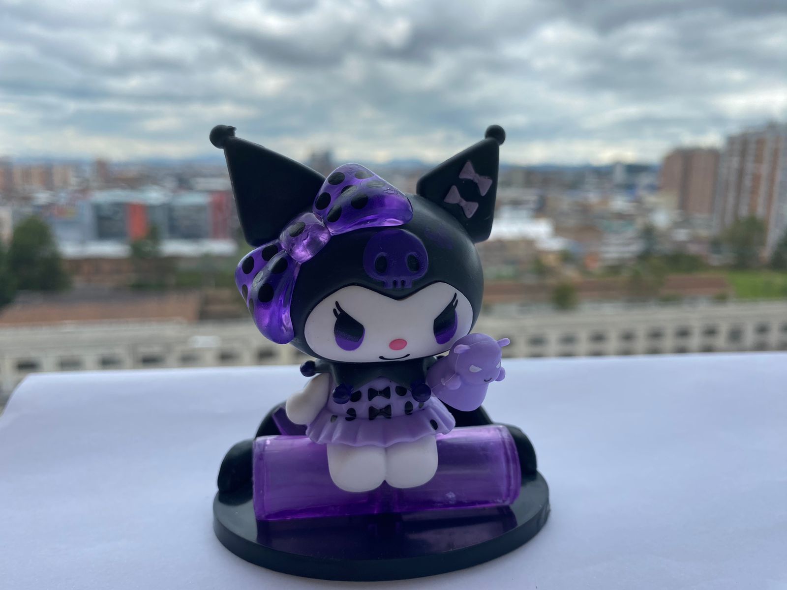 Figura miniatura Kuromi morada con vestido, lazo y accesorio, sobre base negra y morada, fondo urbano nublado.