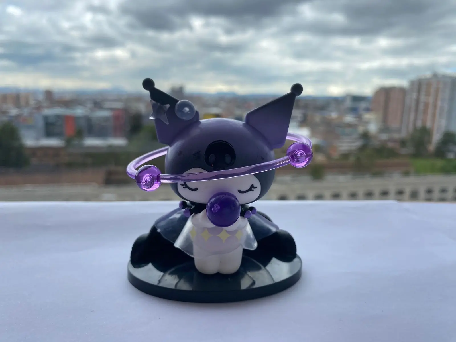 Figura miniatura de Kuromi morada con aro de cuentas púrpuras, sentada sobre base negra, fondo urbano nublado.