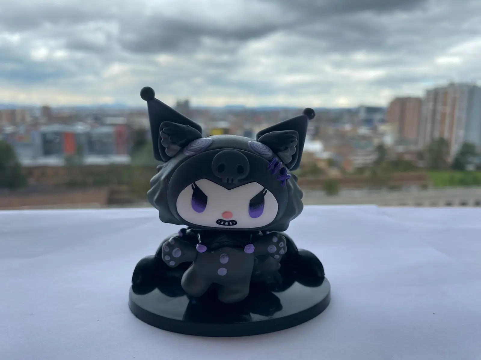 Figura miniatura Kuromi negra con detalles morados, sentada sobre base negra ondulada, fondo urbano nublado.