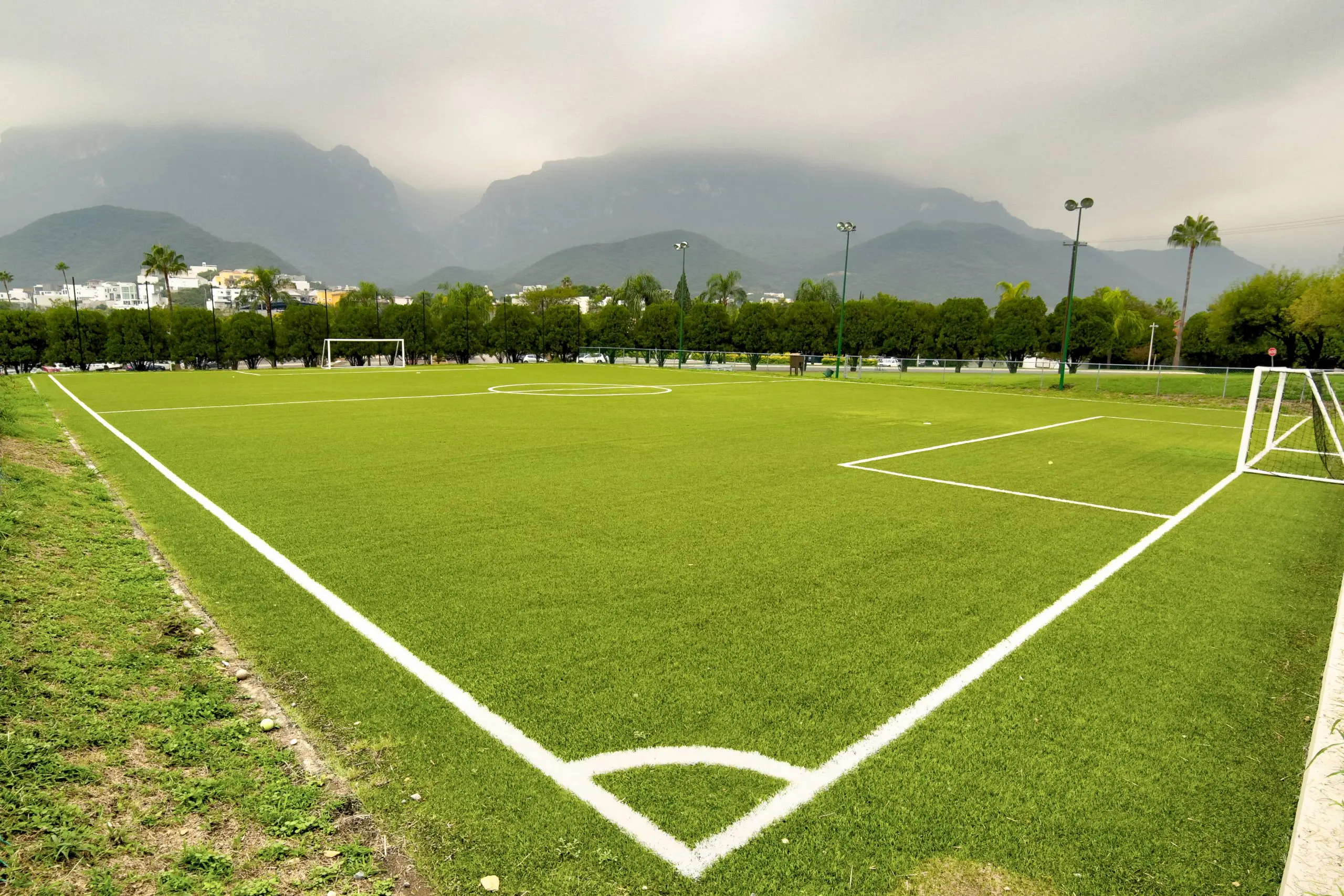 Césped artificial de cancha de fútbol con porterías, montañas neblinosas al fondo y línea blanca de corner.