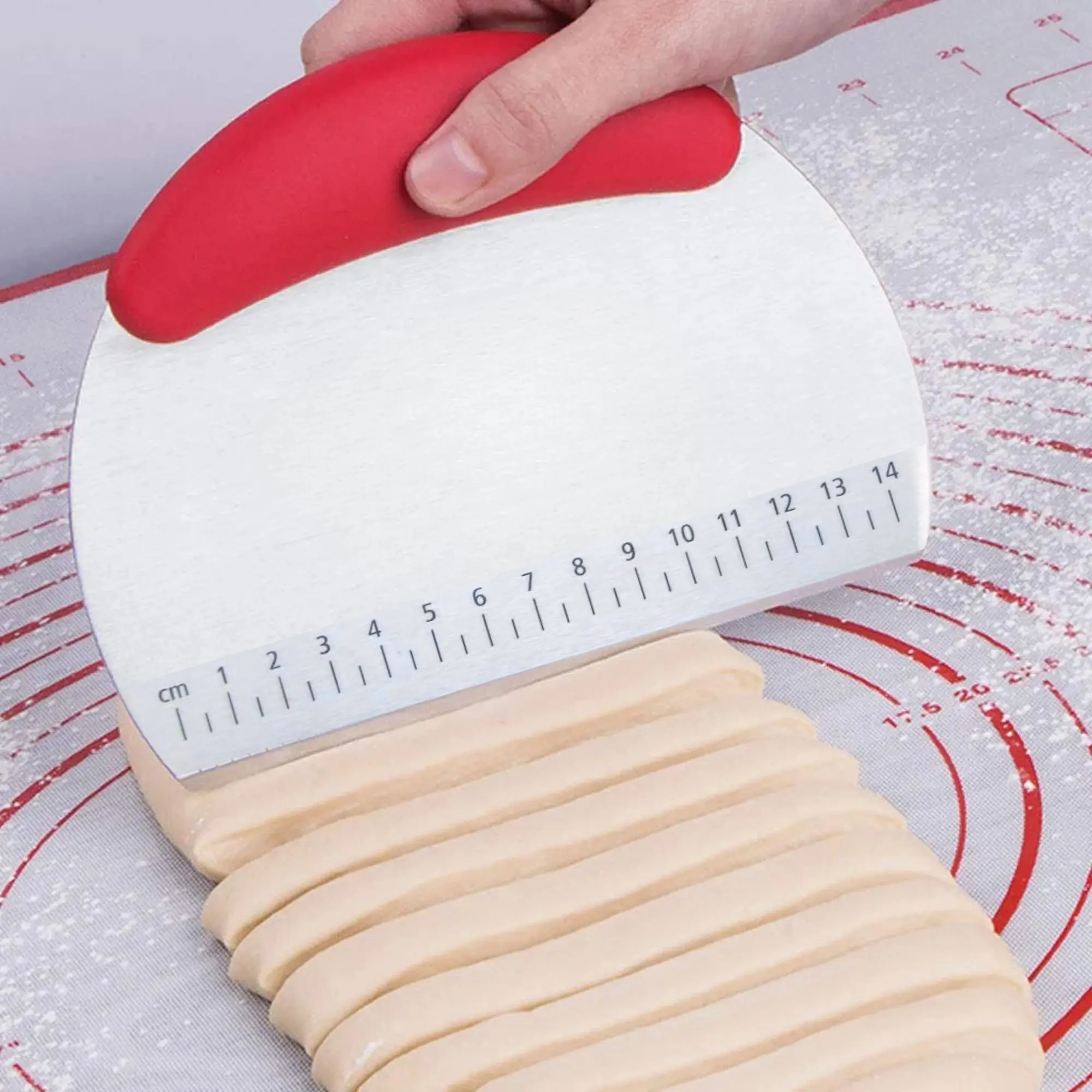 Espátula de cocina de metal con medidor en cm y mango rojo cortando masa en tiras.